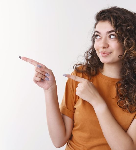 Attractive enthusiastic armenian young woman curly hair pointing looking intrigued left show index fingers awesome blank space advertisement smirking joyfully introducing product, white background.